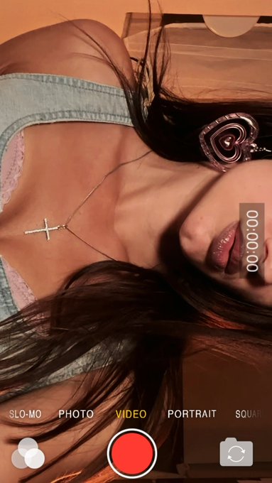 The image shows a woman with long hair wearing earrings, holding her head down while looking at her phone screen. She appears to be taking a selfie or recording something on her device. The photo captures her face and upper body, including her neckline area where she is wearing a necklace. The background of the image is blurred, but it seems to be a room with a bed visible in the distance.