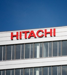 The image shows a corporate building with a prominent red "HITACHI" logo displayed against a clear blue sky. This visual connects to the news that Hitachi is launching a $140 billion initiative to combat Bitcoin and crypto crime by monitoring wallets and suspicious digital-asset flows for banks and crypto firms starting October 2026.