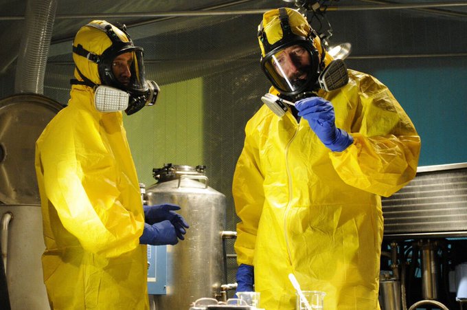 Two figures in yellow hazmat suits and full-face respirators stand in what appears to be a laboratory setting, examining equipment.