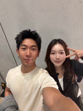 A selfie of a young man in a cream sweater and a young woman in a black jacket making peace signs, taken indoors against a concrete-style wall.