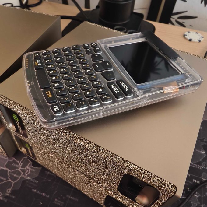 A transparent-cased handheld PDA or pocket computer with a full QWERTY keyboard and small screen sits on top of a cardboard box.