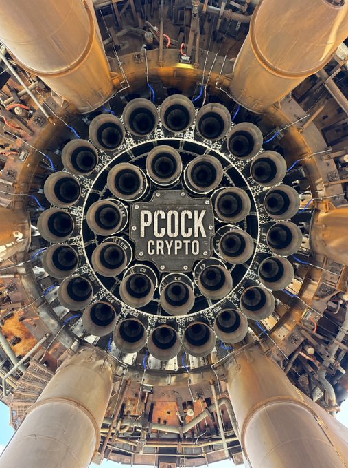 A view of a heavy industrial machine or engine with multiple cylindrical components arranged in a circular pattern, featuring the text "PCOCK CRYPTO" in the center.