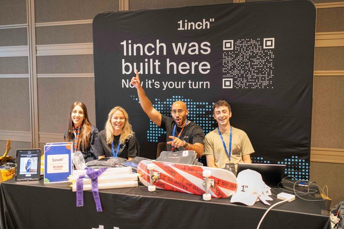 Four people pose energetically at a '1inch' booth under a banner reading '1inch was built here. Now it's your turn' with a QR code.