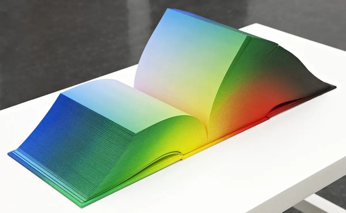 An open art book displayed on a white surface, with its pages fanning upward in a sculptural wave shape showing a rainbow gradient of colors from blue to red.