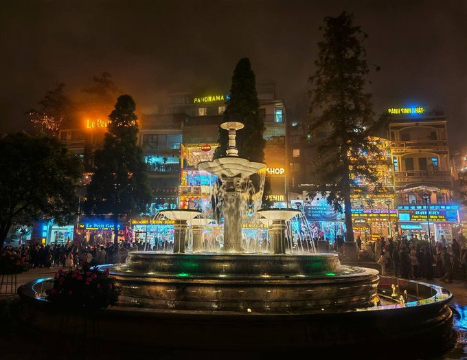 A nighttime cityscape showing an illuminated tiered fountain surrounded by shops with neon signs like 'PANORAMA' and 'PAUL SWISS WATCH'.
