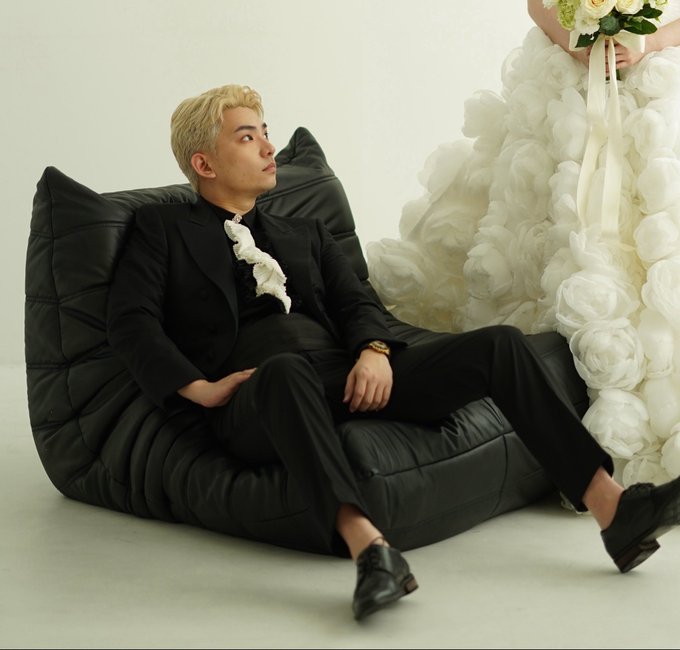 A man in a black suit with blonde hair sits relaxed on a black quilted sofa, gazing upward with a soft expression, while a person beside him—visible only from the torso down—wears a voluminous white gown adorned with large rose-like floral textures and holds a bouquet of white flowers tied with a long ribbon. The minimalist, light-toned backdrop suggests a stylized wedding or pre-wedding photoshoot, with the imagery possibly relating to the post's reference of "accidentally triggered the version answer" through its elegant, almost staged composition.