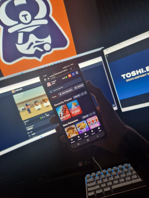 A hand holds a smartphone displaying the TOSHI app store with featured games, set against a backdrop of dual monitors—one showing a TOSHI.BET "200% DEPOSIT BONUS" promo screen—celebrating the partnership announcement with ToshiBet.