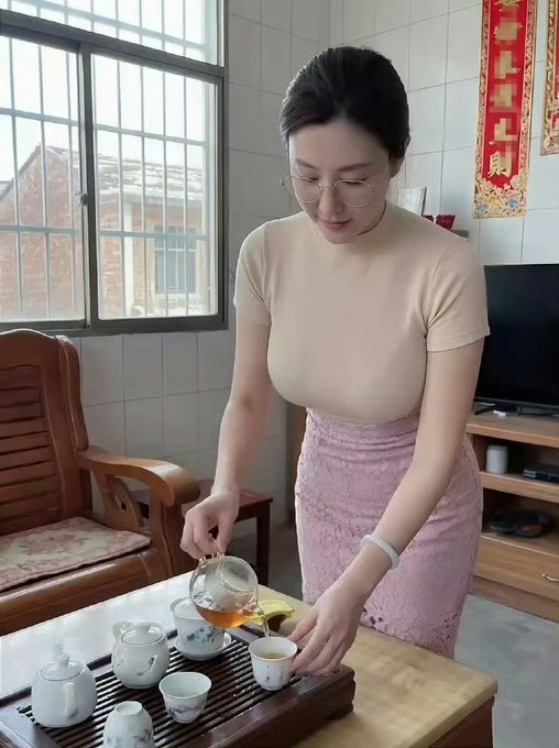 A woman wearing glasses, a beige top, and a pink skirt, pouring tea from a glass teapot into a small cup on a wooden tea table in a living room.