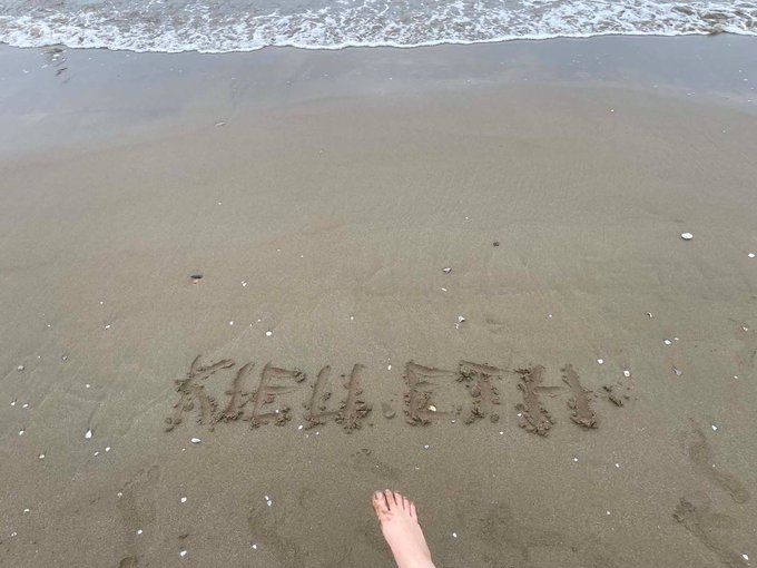 A foot is visible at the bottom of the frame as the words 'HELP ETN' are written in the wet sand at the edge of the ocean.
