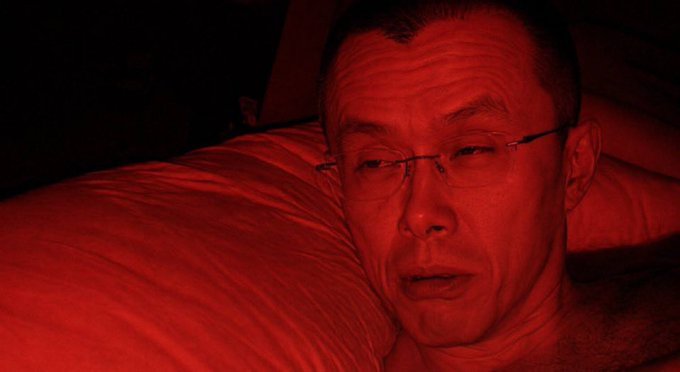A close-up portrait of an older man wearing glasses, his face bathed in deep red lighting as he rests against a pillow.