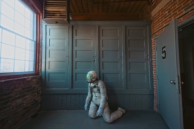 An astronaut in a white spacesuit with a green-reflective visor kneeling on a wooden floor in a rustic room with brick walls, wooden lockers, and a door marked with the number '5'.
