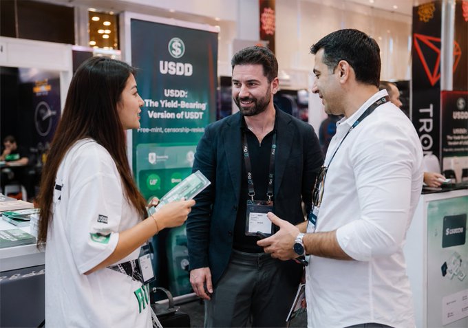 The image shows a group of people gathered around a booth during an industry event or conference. Among them, there are two individuals talking to each other while the others observe. One of these persons is wearing a lanyard that displays their UDI card. The attendees appear interested in discussing various topics related to blockchain and cryptocurrency, as they stand near several banners and displays. These events provide platforms for people from different backgrounds to come together, share ideas, collaborate, and exchange knowledge about the emerging industry.