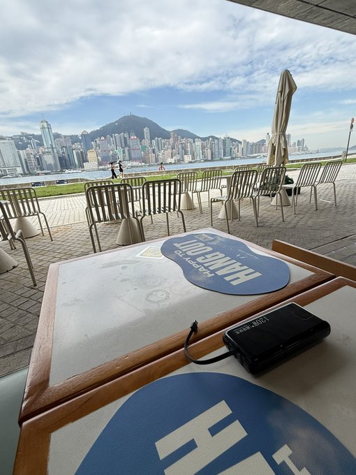 A table with a cardboard 'HK ALL DAY' sign and a pair of KEYS black headphones, overlooking a waterfront view of Hong Kong's cityscape and mountains from an outdoor café area.
