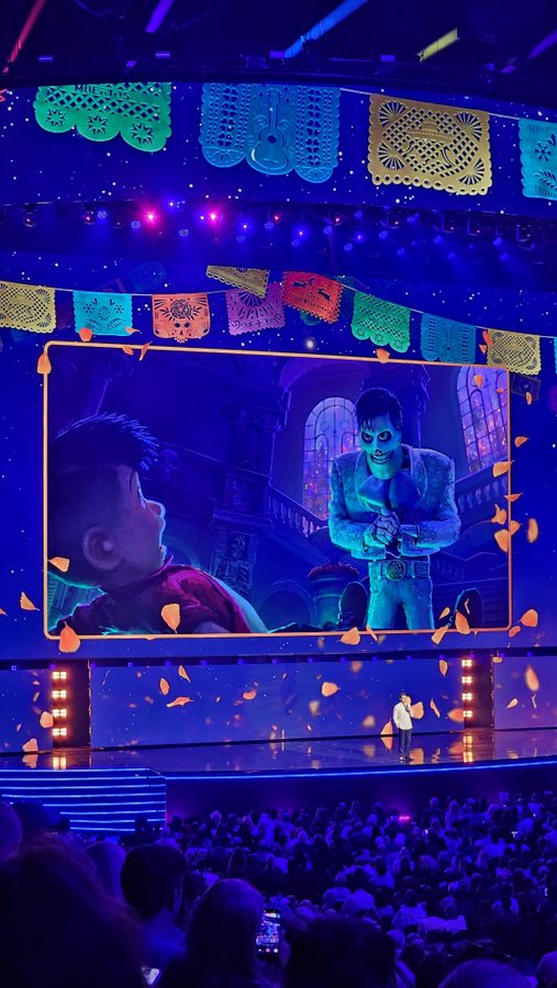 A person stands on a brightly lit stage presenting a scene from Pixar's "Coco" on a large screen, framed by colorful papel picado banners and marigold petals. The caption "me when i have no plot" humorously captures the mood of a presenter sharing an emotional moment from a plot-driven film while lacking their own storyline.