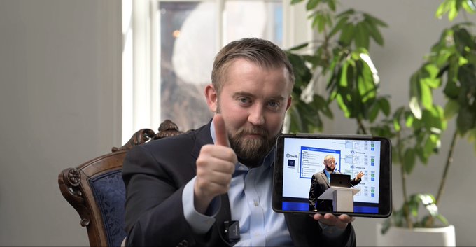 A bearded man in a suit gives a thumbs up while holding a tablet displaying a presentation about 'Personality Insights', seated in a room featuring a portrait.
