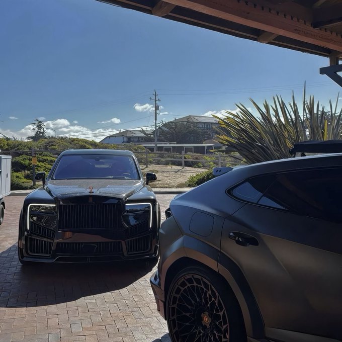 Two luxury SUVs—a black Rolls-Royce and a matte-finished Lamborghini Urus—are parked under a carport on a brick driveway, symbolizing the lifestyle and financial success that effective money management tools aim to provide. The image pairs with the post's message about banking and token utility, using the vehicles as a visual metaphor for financial growth and prosperity.