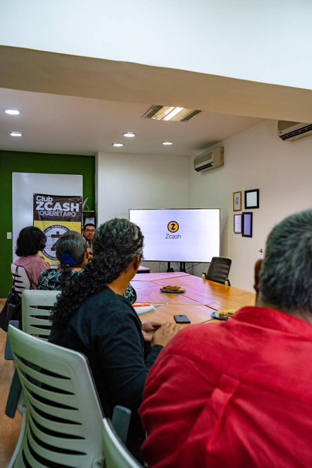 A group of people sit around a large conference table in a meeting room, watching a presentation on a screen displaying a 'Zcash' logo.