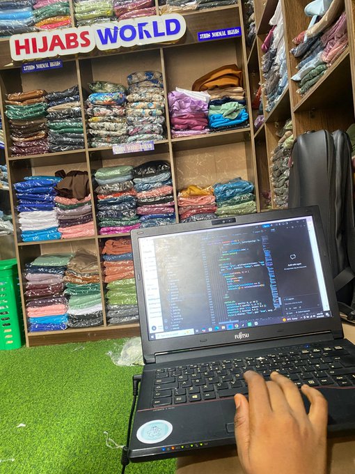 A developer codes on a Fujitsu laptop in a hijab shop, surrounded by neatly folded scarves beneath a "HIJABS WORLD" sign—capturing the "weekend hustle" spirit of grinding code while the shop does business.