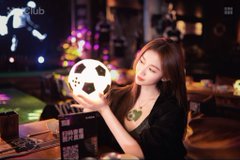A young woman with long brown hair sits in a dimly lit setting, holding up a soccer ball with a black and white pattern near her face.