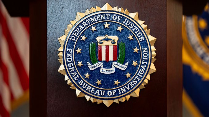 The image shows the official seal of the Federal Bureau of Investigation (FBI), part of the Department of Justice, displayed on a wooden podium with American flags in the background. This emblem relates to the accompanying post about former FBI supervisory agent Patrick Steven Yaroch being charged with allegedly misusing internal bureau systems to steal cryptocurrency linked to an adversarial nation.