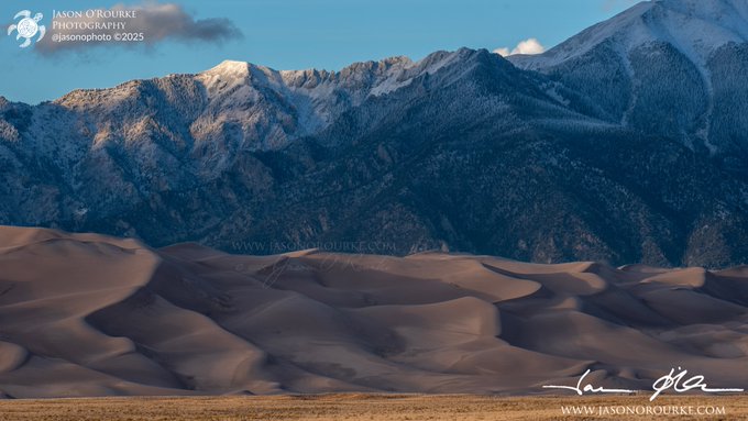 The image features a breathtaking view of snow-capped mountains under a clear blue sky. The mountains are covered with snow, creating a stunning contrast against the sky. In the foreground, there's a large sand dune that adds depth to the scene. The photo is taken from a low angle, making the mountains appear even more majestic and imposing.