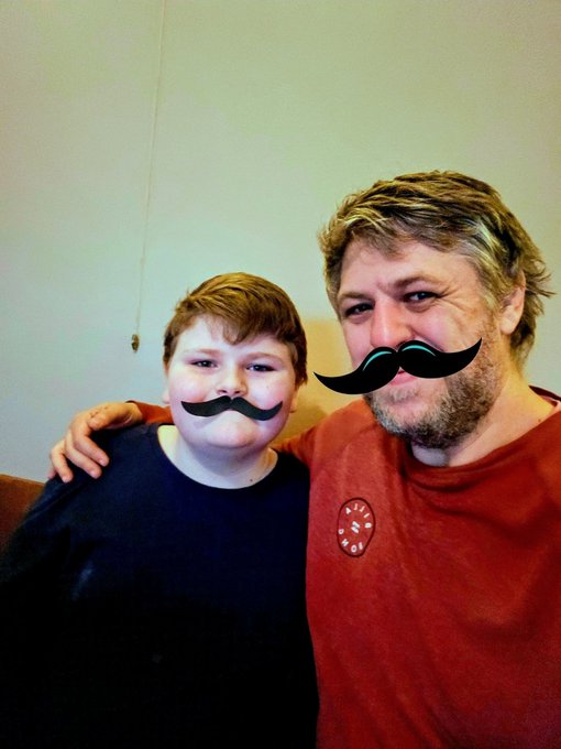 A man and a boy pose together for a photo, with digital black mustache graphics overlaid on their faces. The man wears a red shirt with a circular logo, and the boy wears a dark shirt.