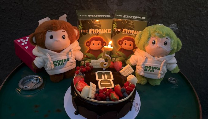 A birthday cake decorated with strawberries and the number '3' candle sits in front of two plush toys labeled 'MONKE SAU', with matching 'THE MONKE' merchandise boxes in the background.
