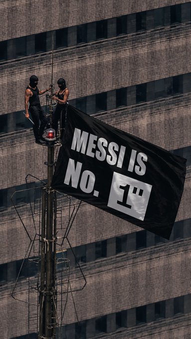 Two masked activists climb atop a communications antenna, unfurling a large black banner reading "MESSI IS NO. 1" in bold white text against the backdrop of a tall brick building.