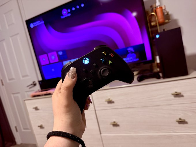 A hand holds up an Xbox controller in front of a TV displaying the Xbox dashboard with a vibrant purple background. The gaming setup on the white dresser reflects the excitement of being ready to play, matching the post's celebration of being "all set up."