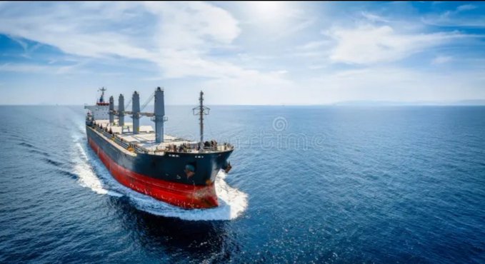 An aerial view of a large red and black cargo ship sailing across the open blue ocean under a partly cloudy sky, with a 'Dreamstime' watermark visible.