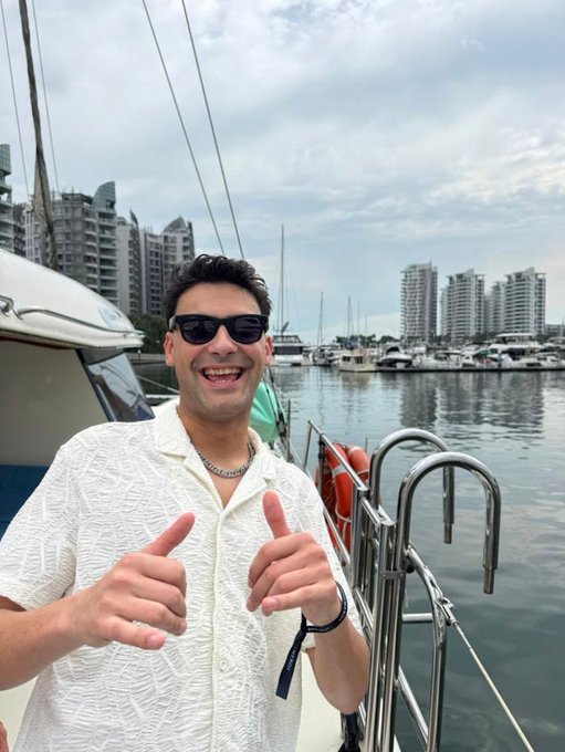 A selfie of a smiling man wearing sunglasses and a white textured shirt on a boat, with a marina and city skyline in the background.