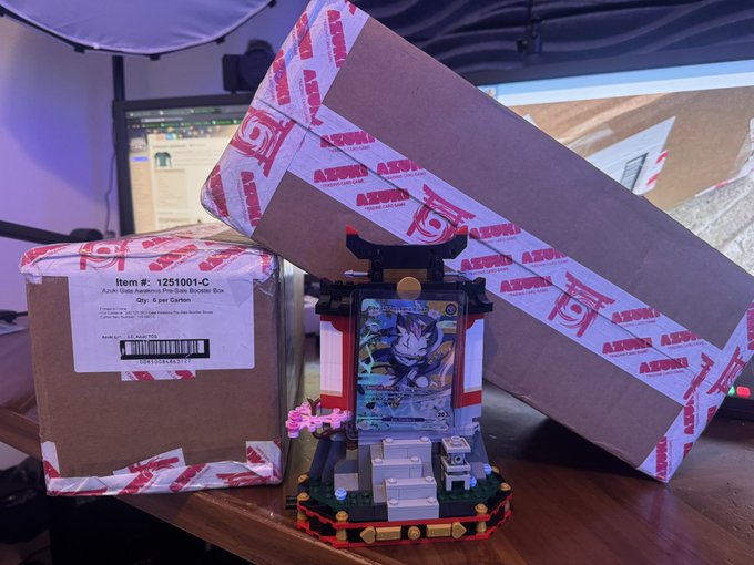 Two Amazon boxes with 'Anime' branded tape sit behind a Naruto-themed LEGO brick display featuring a character and a card, on a desk with monitors in the background.