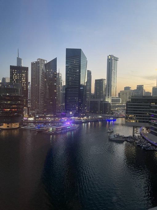 Evening cityscape photograph showing a waterfront marina with high-rise buildings and skyscrapers, likely a modern downtown district at dusk.