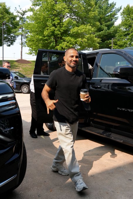 A man in a black t-shirt, light gray sweatpants, and sneakers walks away from a black SUV holding a coffee cup and smiling.
