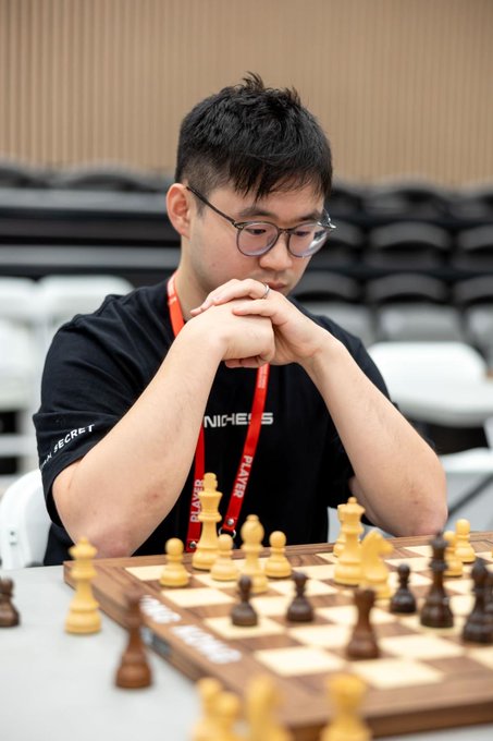 A young man wearing glasses and a black shirt with a red lanyard sits in deep concentration with his hands clasped, playing a game of chess.