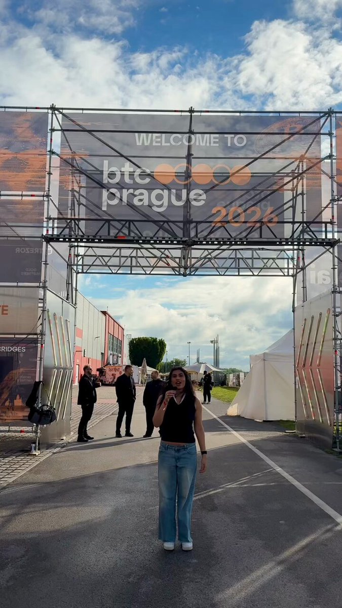 The image features a woman standing outside of an open doors area, with an elaborate metal barricade above her. A "btcprague #prague #pva #bitcoin #letnany" sign is displayed next to the entrance, which seems to be part of the event taking place in Prague or similar locations. The girl appears to be posing for a picture while surrounded by others who may also be attending the gathering.