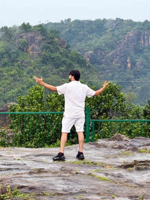 A man in a white shirt and shorts standing on a rocky overlook with arms outstretched, facing a scenic view of forested mountains and rock formations.