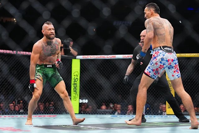 Two MMA fighters face off in a UFC octagon, with the fighter on the left wearing green shorts and the fighter on the right wearing blue and white Venum patterned shorts. A referee stands between them, and visible text includes 'UFC', 'MOBILE ARENA', 'SUPP4KURE.COM', and 'supertec'.