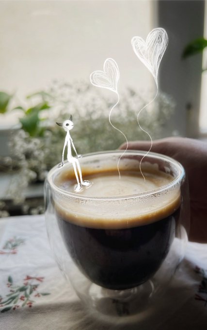 A creative photo of a double-walled glass cup filled with espresso, decorated with a white stick figure sitting on the rim holding strings attached to two heart-shaped balloons.