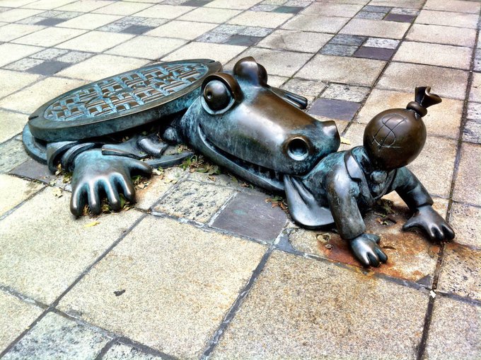 A bronze sculpture depicts a large frog or toad with its mouth open, alongside a smaller humanoid frog character with a grenade-shaped head, set into a tiled pavement.