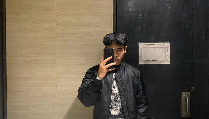 Mirror selfie of a young man with dark hair wearing a black jacket and skull-print shirt, holding a phone.