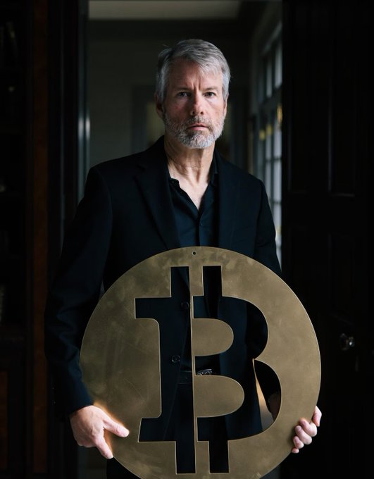 A man with grey hair and a beard, wearing a black suit, stands in a doorway holding a large brass Bitcoin symbol with the 'B' cut out.