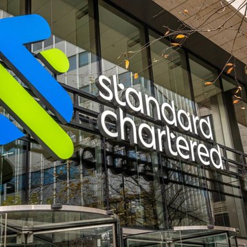 The image shows the exterior of a glass building with Standard Chartered's logo displayed prominently above a circular window entrance. The logo consists of a blue and green square, possibly representing the bank's name or initials. A text below the logo mentions Bitcoin's current low price as being "almost in" and could signify the bank considering buying Bitcoin ETF holdings to enter this market segment. Furthermore, it references Strategy's expected buyback which suggests that the bank might be closely monitoring its investments in digital assets like Bitcoin. The text is located close to the viewer and appears to be the focal point of the image.