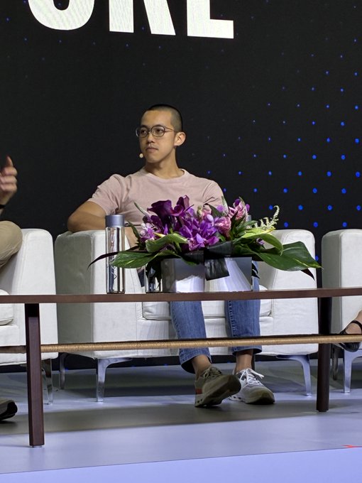 A young man wearing a pink shirt and jeans sits on a white couch on a stage with a starry background, with a coffee table and a bouquet of purple flowers in front of him.