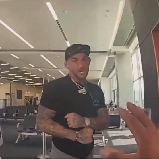 A muscular man in a black t-shirt, backwards cap, and silver chain stands in what appears to be an airport terminal, gesturing with his arms while talking to someone off-camera.