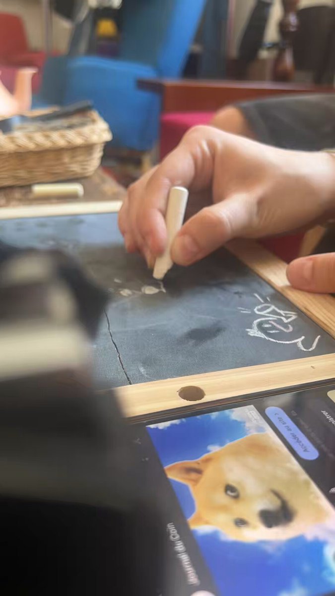 The image shows a person's hand holding a white marker, drawing on a blackboard or chalkboard with a picture of a dog's face. The scene takes place at a desk where there are two cell phones visible - one placed near the bottom right corner and another towards the top left corner. A chair is also present in the background, positioned behind the person who is drawing. The image includes text that reads "When I ask to my Intern to draw Kabosu head because my business accept Dogecoin To the moon".
