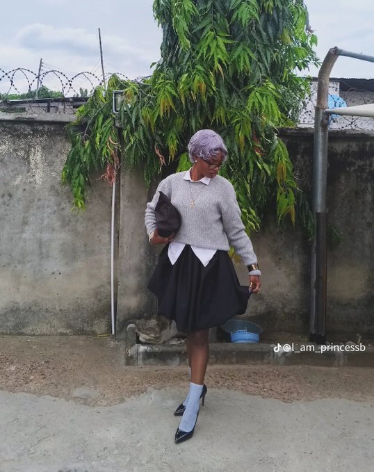 The image features a woman wearing a gray sweater, black skirt, and black high heels while standing outside of a building with a fence behind her. She has a purse hanging from her shoulder and appears to be posing for the camera. The photo is titled "Lamam princesseb" in the caption, which suggests that it might have been taken by someone named Lamam or a similar name who posted the image on social media platforms like Facebook, Instagram, or Twitter.