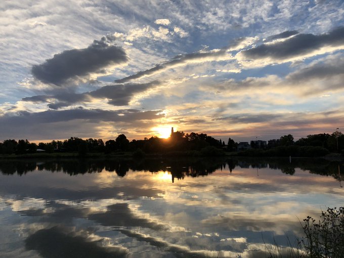 A scenic landscape photo of a sunset over a lake, with the sky and clouds reflected in the calm water and a silhouetted treeline on the horizon.