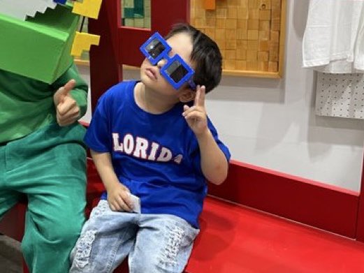 The image features a young boy sitting on a red bench, wearing a blue shirt with "lboro" written on it. The boy has his eyes closed and is making a peace sign with his fingers. In front of him, there are two other people who appear to be dressed in green and white clothing. They seem to be posing for the photo or observing something interesting nearby.