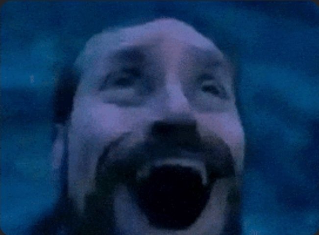 A heavily distorted, low-resolution image of a bearded man with his mouth open, tinted in dark blue tones, appearing like a glitchy frame from a movie.